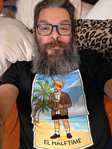 Reviewer lying down for a selfie in the black El Halftime OG T-Shirt, showing the Lotería-style graphic.