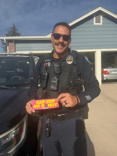 Reviewer in uniform smiling while holding a pink "BAD COP NO DONUT" sticker from the TCG92 MYSTERY CASH BOX.