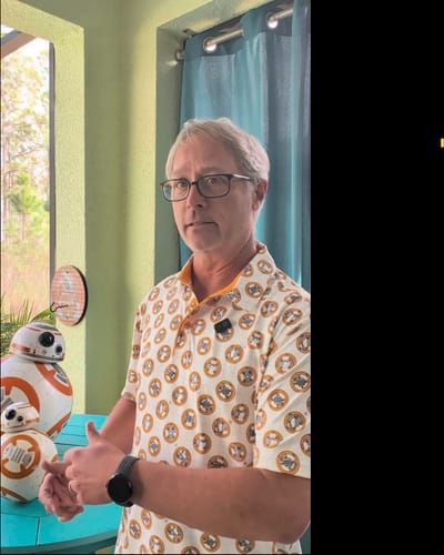 Partial view of a reviewer wearing the patterned 'The BB-8' – All-Day Polo, standing next to two BB-8 droid figures.