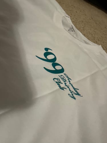 Close-up of a customer's white MYSTERY TEE, showing the teal '99 Tuesday Driving Club' logo on the chest.