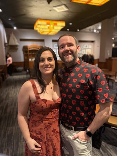 Reviewer smiling while wearing the black Disney "Lovebirds" shirt with red hearts, standing next to a companion indoors.