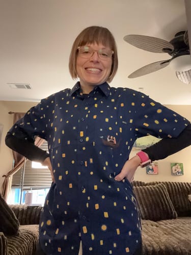 A reviewer smiles while wearing the navy Disney's Tangled "Lanterns" short sleeve shirt, layered over a black long-sleeve top.