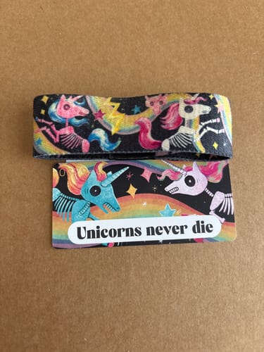 Reviewer's 'Unicorns Never Die' bracelet and card showing colorful skeletal unicorns against a dark background.