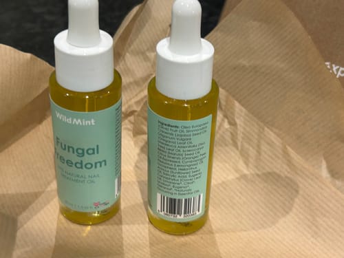 Reviewer showing the front and back of two Fungal Freedom nail oil bottles, resting on packaging paper.
