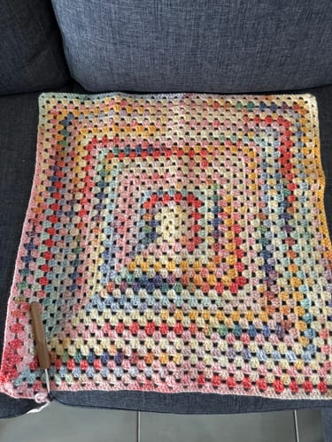 Customer's Hearthside Rainbow Blanket in progress. The colorful, crocheted square is laid on a sofa with the hook attached.