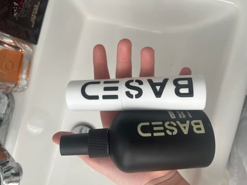 Customer's hand holding a black bottle of BASED Texture Powder and a white branded tube over a sink.