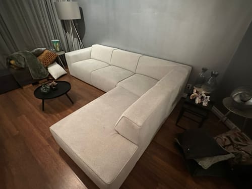 Recensent toont de naturelkleurige by fonQ Stretch hoekbank in een woonkamer.