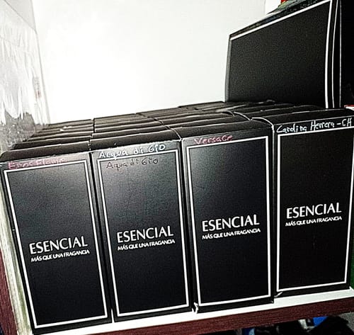 El cliente muestra su pedido de perfumes Esencial, con varias cajas negras alineadas en una estantería.