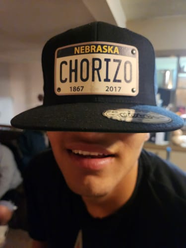 A smiling person wears the black Nebraska Plate Hat with a custom 'CHORIZO' license plate patch pulled low over their eyes.
