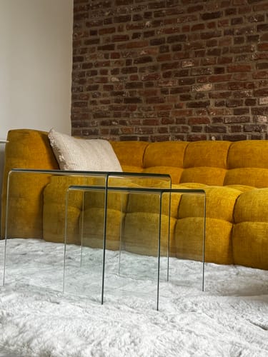 Beoordelaar toont de transparante Vetro bijzettafel set op een pluizig vloerkleed voor een gele bank.