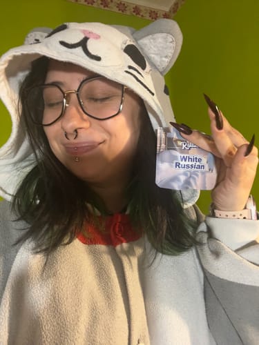 Reviewer in a fun cat hoodie smiles while holding up the White Russian THCA Flower packaging.