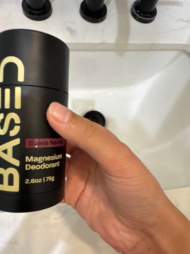 A customer's hand holds a black tube of BASED Deodorant in the Guava Nectar scent over a bathroom sink.