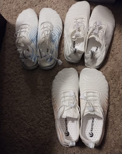 Reviewer's three pairs of white Lorax Pro barefoot shoes with wide toe boxes arranged on a carpet.