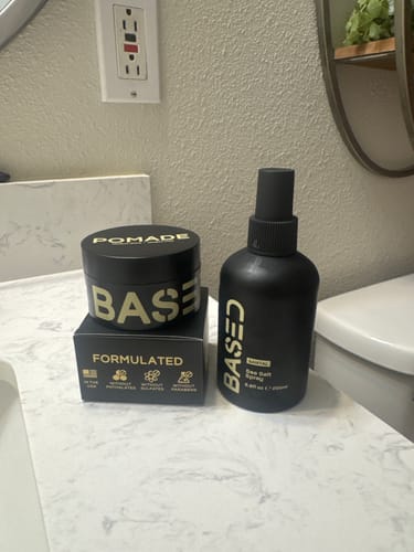 Customer's black jar of Based Pomade and bottle of sea salt spray sitting on a bathroom counter.