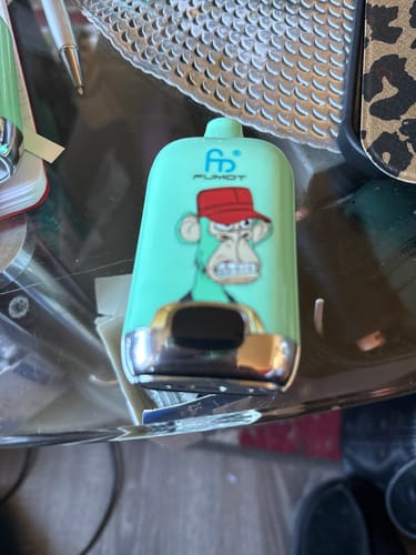 Ein Kunde zeigt die hellgrüne RANDM DIGITAL BOX 12000 Vape mit Cartoon-Affen-Aufdruck auf einem Tisch.