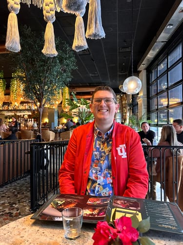 Reviewer smiling at a restaurant table while wearing the Disney's The Muppets "Picture Perfect" shirt under a red jacket.