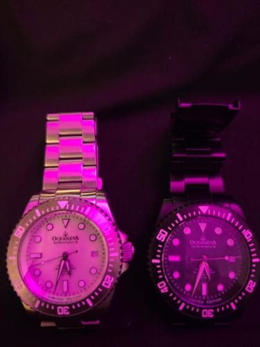 Customer's silver Oceaneva Deep Marine Explorer III watch next to a black model, both with glowing lume under a purple light.
