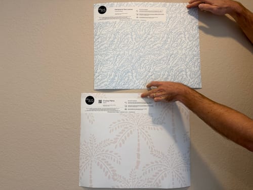 Customer holding a sample of the Hamptons Sea Leaves in Coastal Blue wallpaper against a neutral wall.