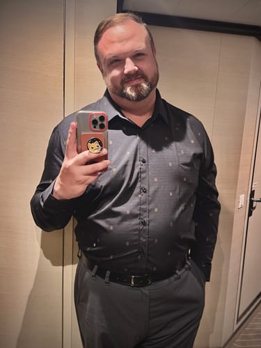 Reviewer taking a mirror selfie while wearing the dark, patterned Arch Deco KUNUFLEX dress shirt tucked into grey trousers.