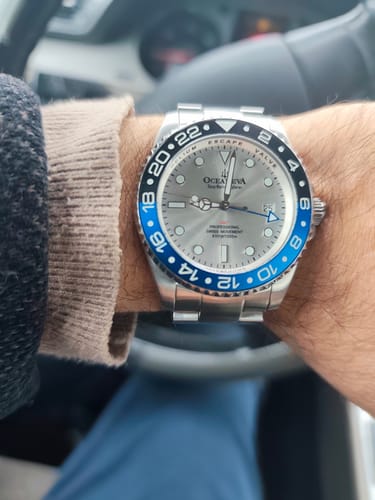 A reviewer's close-up of the Oceaneva GMT Deep Marine Explorer watch on their wrist, showing the silver dial and blue/black bezel.
