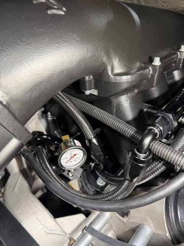 Close-up of the reviewer's Fluidworks regulator bracket installed in an engine with a pressure gauge and black braided hoses.