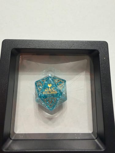 Reviewer's personalized blue D20 dice with gold details, presented in a black shadow box display case.