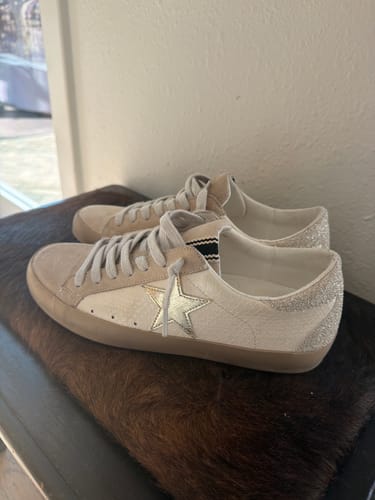 Customer's off-white sneakers with a gold star, suede trim, and a silver glitter heel, shown resting on a furry surface.