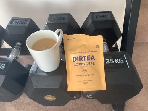 Reviewer's pouch of Cordyceps Mushroom Powder and a prepared drink in a mug, resting on a rack of black dumbbells.