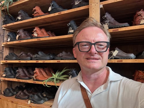 Reviewer smiling in a store with wooden shelves displaying various Karoo Clogs in brown and black.