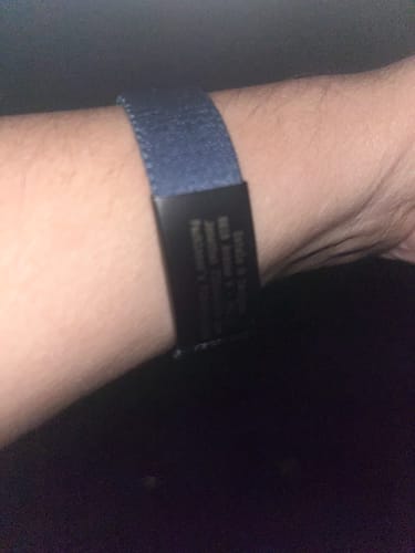 Close-up of a reviewer's wrist, showing the black Medical Alert Bracelet Charm on a dark blue wristband in a dim setting.