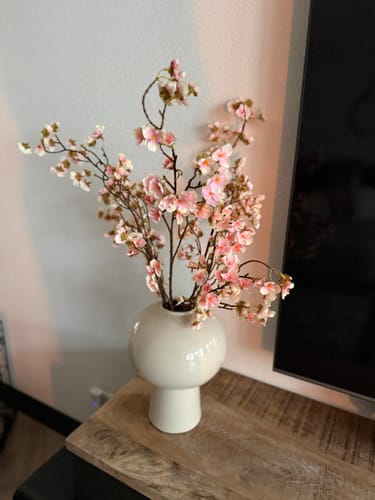 Klant toont de mooie, oudroze Zijden Bloesem takken in een witte bolvaas op een houten meubel.