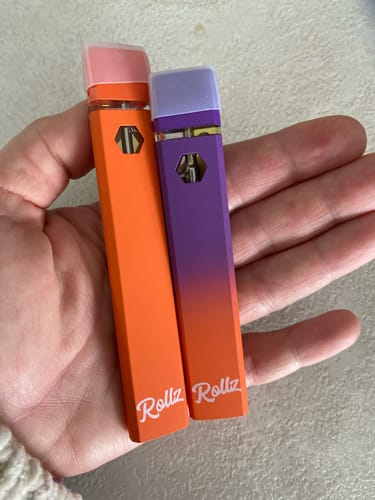 Reviewer holding two 10-OH Superior LIVE RESIN VAPES, one orange and one purple, in their palm.