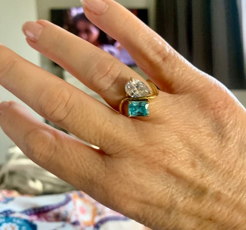 Customer's hand wearing the Navae Birthstone Ring with its clear pear-cut and blue radiant-cut stones on a gold band.