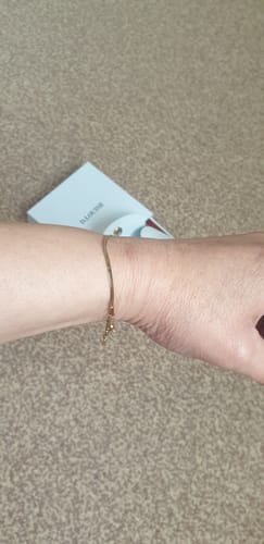 Customer wearing the thin, gold Fine Essentials Bracelet on their wrist, with the product box in the background.