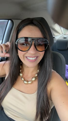 Avaliadora sorrindo em uma selfie no carro, usando os grandes Óculos Veneto pretos.