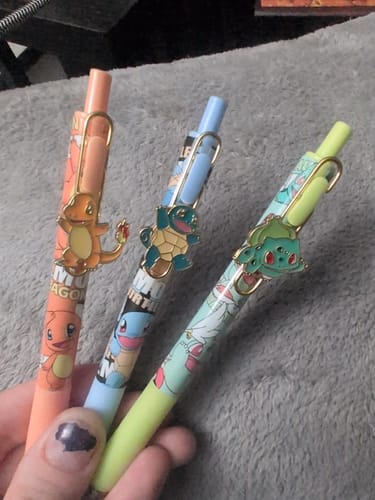 Customer holding three Pokemon gel pens: an orange Charmander, a blue Squirtle, and a green Bulbasaur.