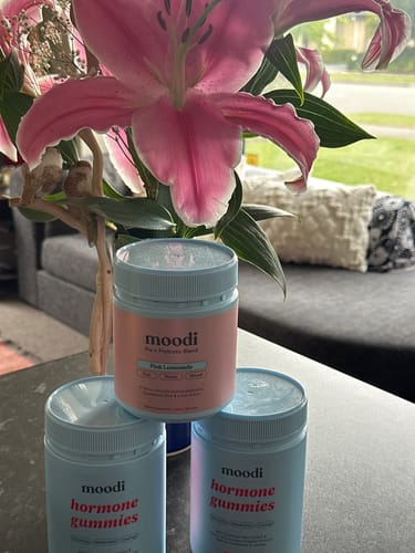 Customer's three bottles of Hormone Gummies stacked in a pyramid arrangement with a large pink lily in the background.