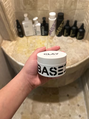 Customer holding the white container of Based Bodyworks Hair Clay in a bathroom with other products on a shelf.