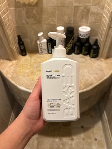 Customer holding the white Based Body Lotion bottle in a tiled bathroom.