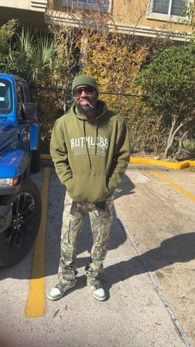 Reviewer standing outdoors wearing an olive green Ruthless Hustle Hoodie with hands in the front pocket.