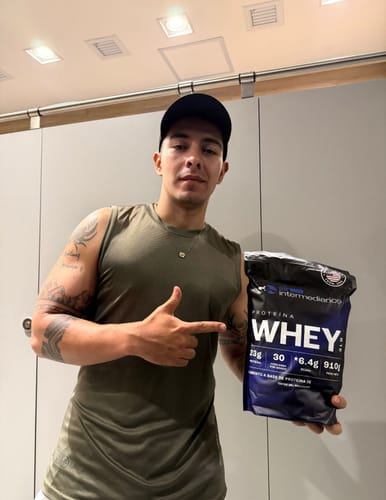 Cliente con camiseta verde sin mangas y gorra negra, señalando una bolsa de Proteína Whey que sostiene en la mano.