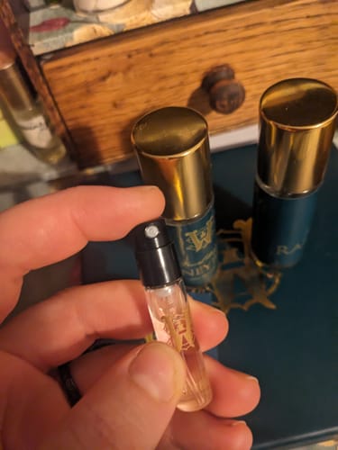 Reviewer holding a small sample vial of Honeyberry perfume with larger bottles in the background.