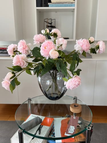 Beoordelaar toont de roze Zijden Pioenrozen in een donkere glazen vaas op een glazen salontafel.