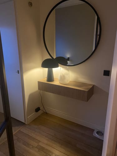 Recensent toont de houten Jolie sidetable die aan de muur hangt met een lamp en vaas erop.