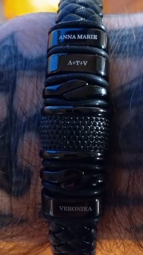 Close-up of a customer's personalized black bracelet on their wrist, engraved with the names 'Anna Marie', 'A+T+V', and 'Veronika'.