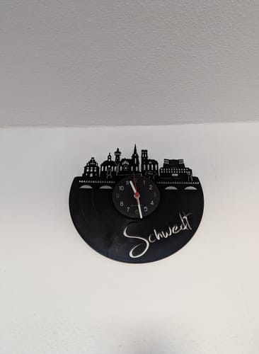 Ein Kunde zeigt die schwarze Vinyl-Wanduhr "Skyline Schwedt", die an einer weißen Wand hängt.