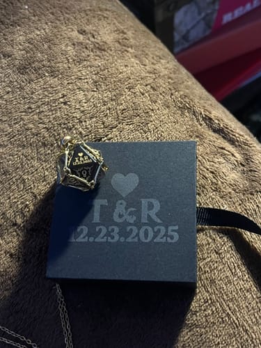 Customer's dark D20 dice pendant resting on a personalized gift box engraved with the initials 'T & R' and a date.
