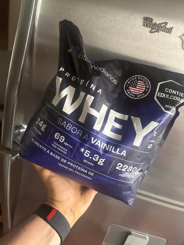 Cliente sosteniendo el empaque grande de Proteína Whey sabor Vainilla frente a un refrigerador.