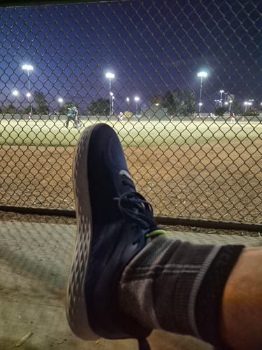 Reviewer's foot in a dark blue athletic shoe resting near a fence, with a night baseball game in the background.