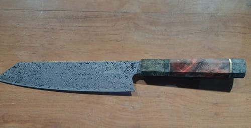 Rezensent zeigt das Damast Gyuto Messer, das flach auf einer Holzoberfläche liegt und seine gemusterte Klinge und den Holzgriff zeigt.